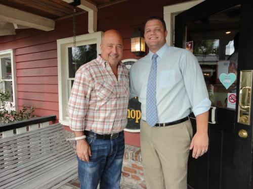 Two men smiling together outside a restaurant, one in a plaid shirt and the other in a dress shirt and tie.
