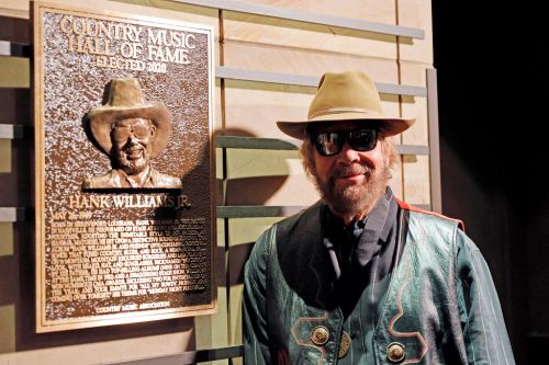 Hank Williams Jr. stands beside his Country Music Hall of Fame plaque, wearing sunglasses and a green vest.
