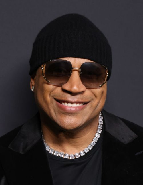 A smiling man wearing sunglasses, a black beanie, and a sparkling necklace, against a dark background.