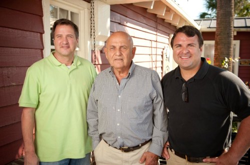 Three men stand together outside a house, smiling at the camera. The background features a red wooden exterior.