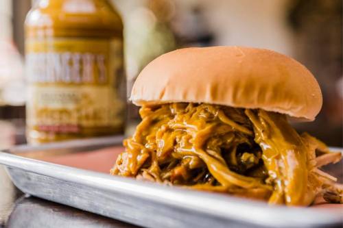 A close-up of a pulled pork sandwich with melted cheese on a soft bun, served on a tray with a bottle of sauce in the background.