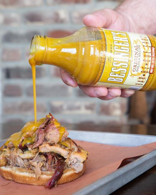 A hand pours yellow sauce over a pulled pork sandwich on a wooden table.
