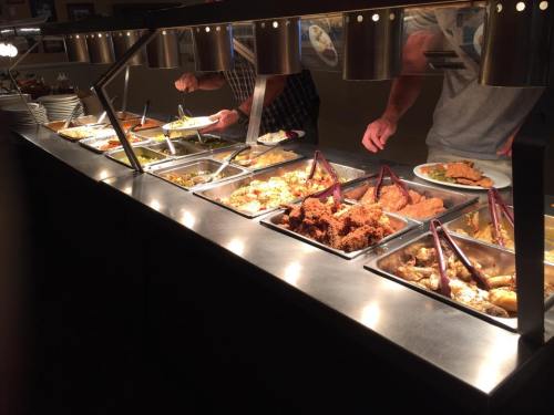 A buffet with various dishes, including fried chicken, being served by diners using tongs.