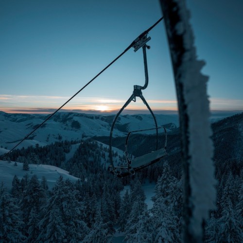 A ski lift chair against a snowy landscape at sunset, with mountains and trees in the background.