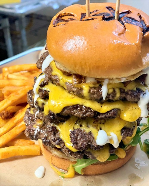 A towering burger with multiple beef patties, melted cheese, and sauce, served with a side of golden fries.