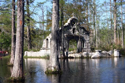 Ruins of a stone structure surrounded by water and cypress trees in a serene, natural setting.