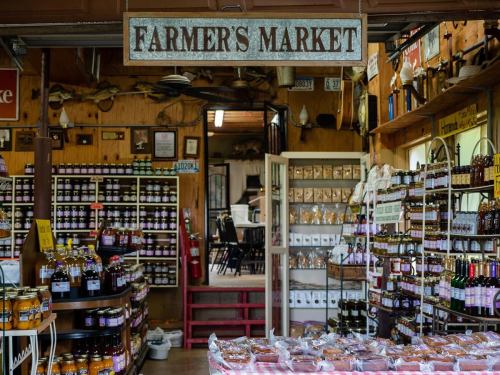 Interior of a farmer's market with shelves of jars, bottles, and packaged goods, featuring a rustic wooden sign.