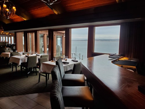 A restaurant interior with elegant tables and chairs, featuring large windows overlooking a calm sea.