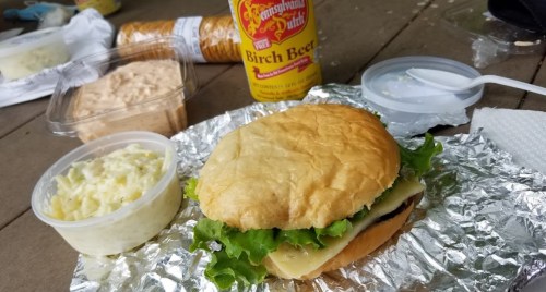 A sandwich with lettuce and cheese on foil, accompanied by coleslaw, a dip, and a bottle of birch beer.