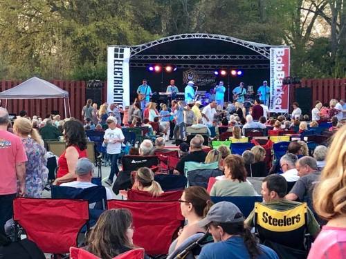 A lively outdoor concert scene with a crowd enjoying music, seated in folding chairs, and a band performing on stage.
