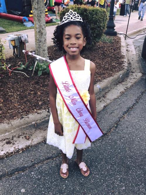 A young girl in a yellow dress and tiara smiles, wearing a sash that reads "Little Miss Grass Roots."