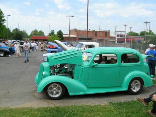 A vintage turquoise car with an open hood, surrounded by people and classic cars at an outdoor event.