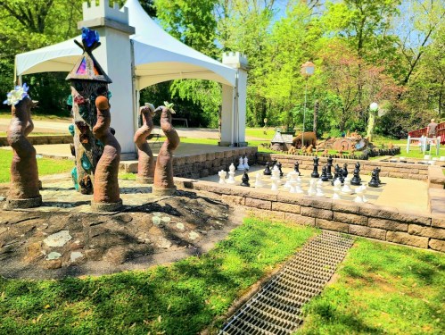 A park scene featuring a large outdoor chessboard and artistic sculptures surrounded by greenery.