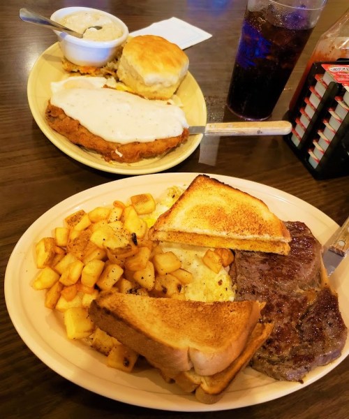A plate of fried chicken with gravy, biscuits, and a side of potatoes, alongside a plate of steak and toast.
