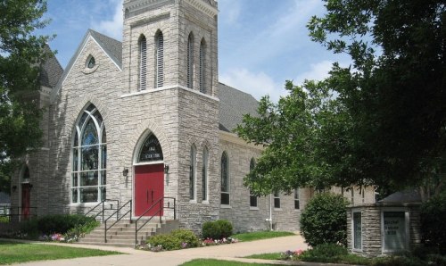 A stone church with a tall steeple, red doors, and large windows, surrounded by greenery and flowers.