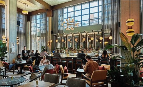A stylish restaurant interior with large windows, a bar, and patrons dining at tables amidst modern decor and plants.