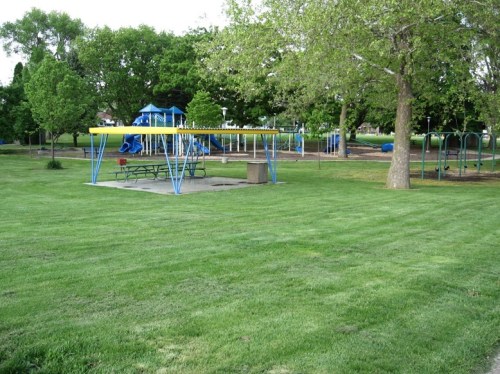 A grassy park featuring a playground, swings, and picnic area under trees on a sunny day.