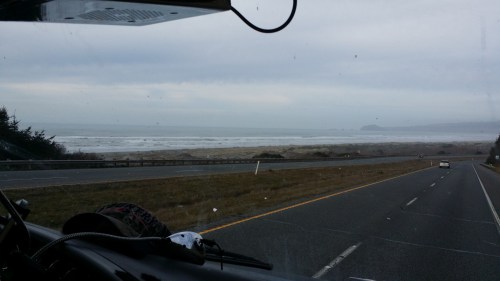 View from a vehicle showing a coastal highway with ocean waves and a cloudy sky in the background.