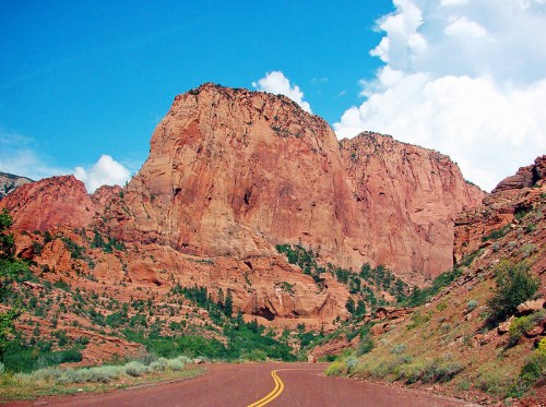 A winding road leads through a canyon with towering red rock formations under a blue sky with fluffy clouds.