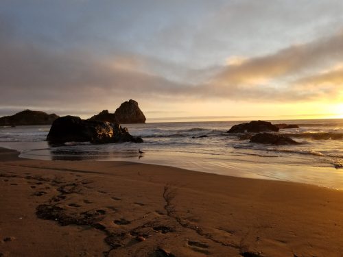 A serene beach at sunset, with gentle waves, rocks in the water, and a cloudy sky reflecting warm colors.