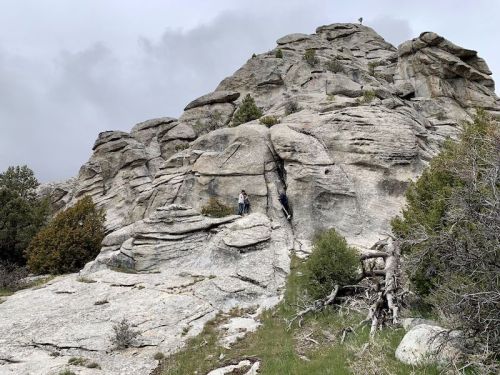 Two hikers navigate a rocky mountain trail under a cloudy sky, surrounded by sparse trees and rugged terrain.