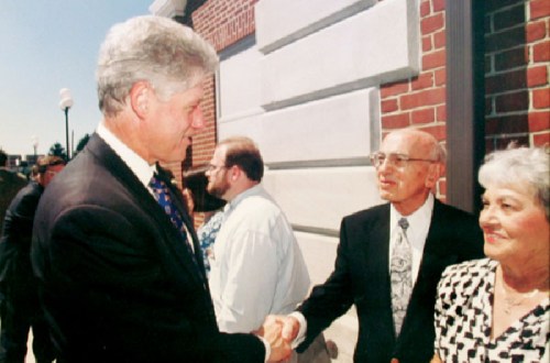 Former President Bill Clinton shakes hands with a man while a woman looks on, with others in the background.
