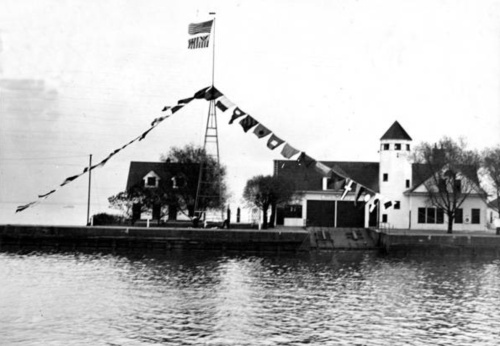 Historic waterfront scene featuring a building adorned with flags and an American flag on a pole, reflected in the water.