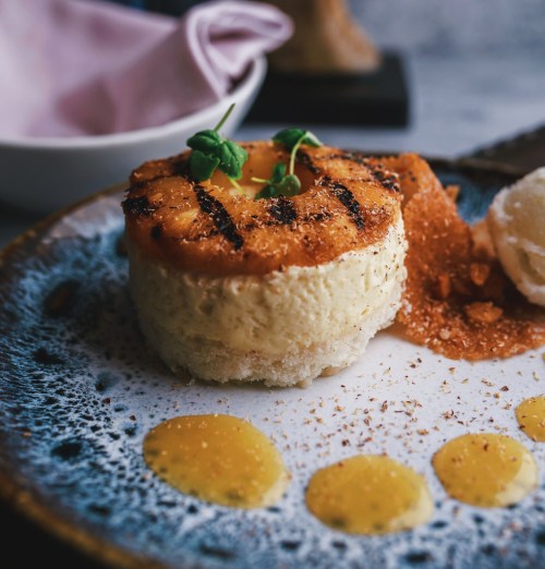 A creamy dessert topped with grilled pineapple, garnished with green herbs and a sprinkle of crumbs on a plate.