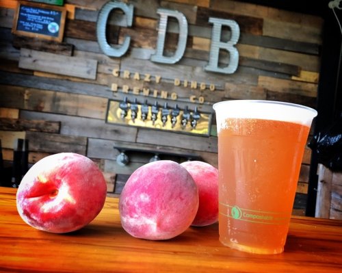 A clear cup of peach beer sits on a wooden table next to fresh peaches, with a brewery logo in the background.