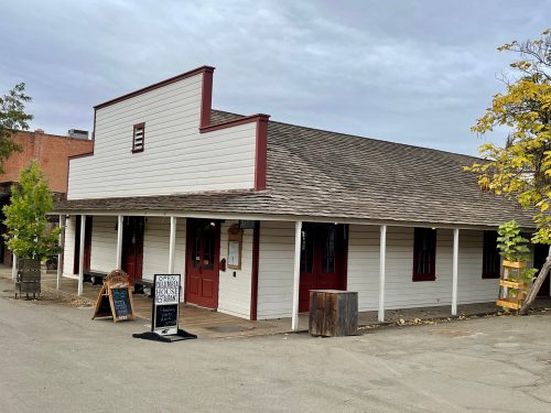 A quaint, white wooden building with red trim, featuring a porch and signs outside, set in a rustic area.