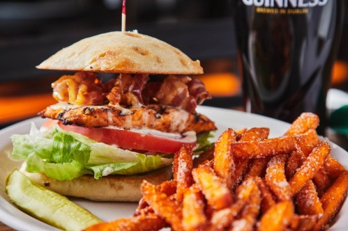 A grilled chicken sandwich with bacon, lettuce, and tomato, served with sweet potato fries and a pickle on the side.