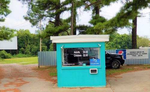 A turquoise ticket booth with prices for adults and children, surrounded by trees and a drive-in movie screen in the background.