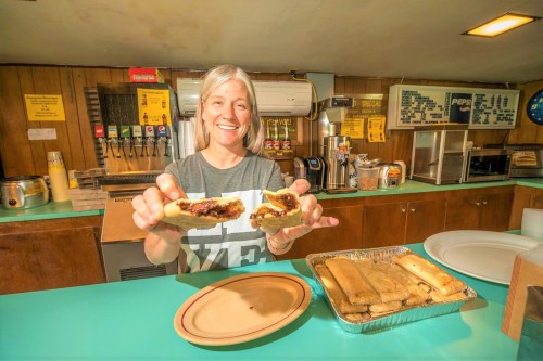 A smiling woman holds two tamales in a kitchen, with a tray of tamales and drink dispensers in the background.