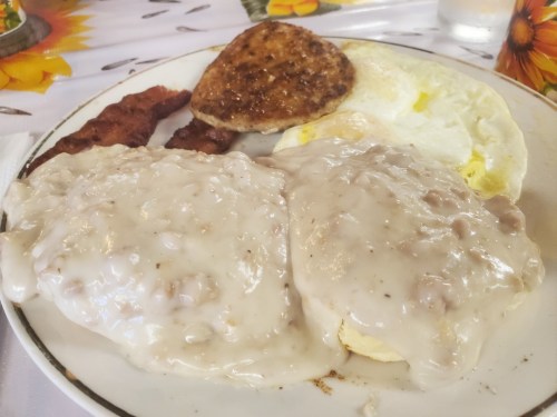 A plate of breakfast featuring biscuits and gravy, sausage patty, bacon, and a fried egg.