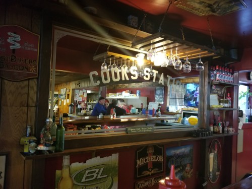 A cozy bar interior with a mirrored counter, hanging glasses, and a glimpse of patrons in a warm, inviting atmosphere.