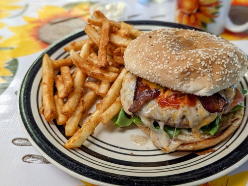 A sesame seed bun burger with cheese, bacon, and lettuce, served with crispy French fries on a patterned tablecloth.