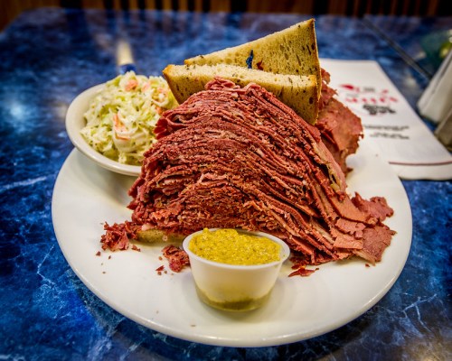 A plate featuring a large stack of pastrami, two slices of rye bread, coleslaw, and a small cup of mustard.