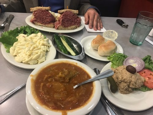 A table set with a plate of sandwiches, macaroni salad, pickles, rolls, and a bowl of soup.