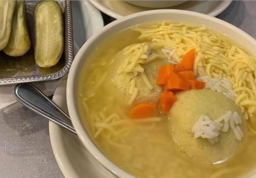 A bowl of soup with dumplings, carrots, and noodles, accompanied by a side of pickles on a silver tray.