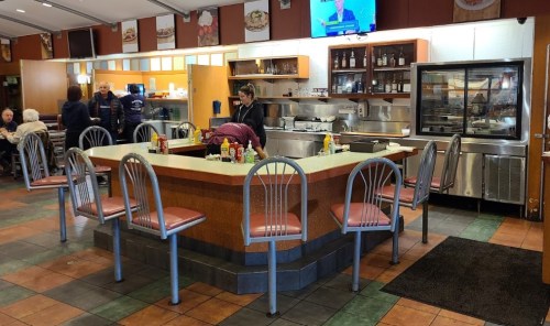 A diner interior featuring a central counter with chairs, condiments, and a kitchen area in the background.