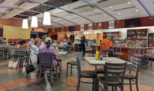 A busy diner with patrons seated at tables, a counter in the background, and bright overhead lighting.