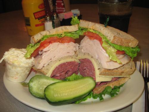 A hearty sandwich with turkey, roast beef, lettuce, and tomato on multigrain bread, served with coleslaw and a pickle.