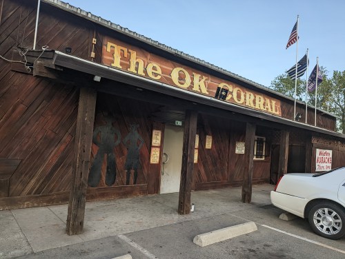 Exterior of The OK Corral, a wooden building with a sign, flags, and a parking lot.
