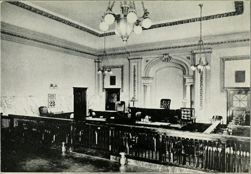 Historic courtroom interior with wooden benches, a judge's bench, and ornate lighting fixtures.