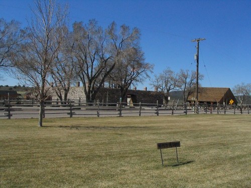 A grassy area with a sign, trees, and rustic buildings under a clear blue sky. Fencing lines the property.