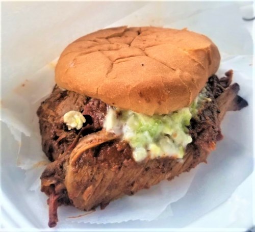 A pulled pork sandwich with coleslaw on a soft bun, served on a white paper plate.