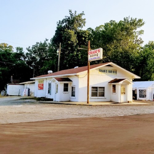 A small, rustic barbecue restaurant with a sign reading "Craig's Bar-B-Q," surrounded by trees and gravel.