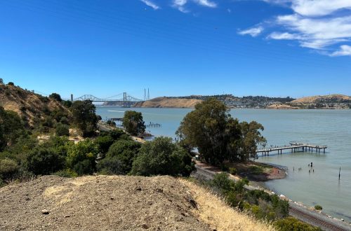 Scenic view of a river with a bridge in the background, surrounded by hills and greenery under a clear blue sky.