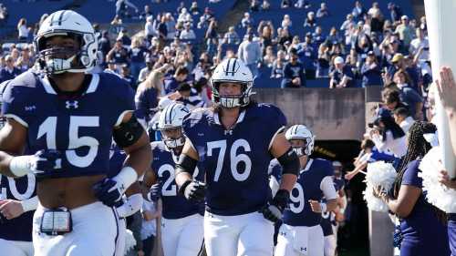 A group of football players in blue uniforms run onto the field, greeted by cheering fans in the stands.