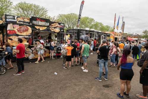 A busy food truck festival with people in line for various food vendors under a cloudy sky.